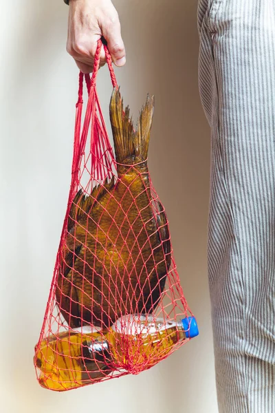 woven mesh bag with a large dried fish and a bottle of beer in his hand man in white trousers with a fine vertical strip