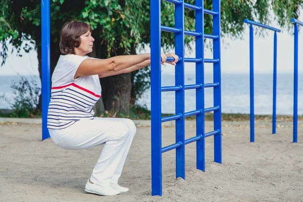 elderly woman performs squatting on summer sports ground