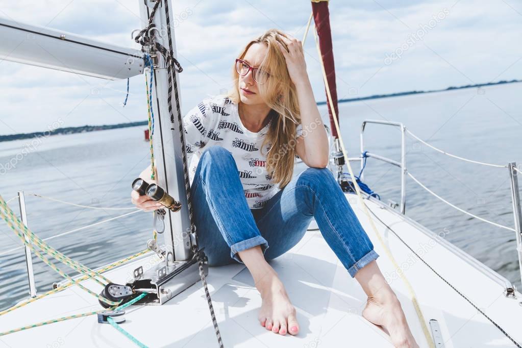 slender barefooted blonde in spectacles sits on nose of yacht