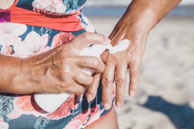 Elderly woman in swimsuit using cream for hand on beach clipart