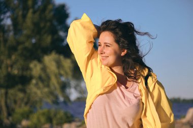 Geniş bir nehrin kumlu sahilinde uzun kızıl saçlı ve parlak sarı ceketli güzel bir kızın portresi.