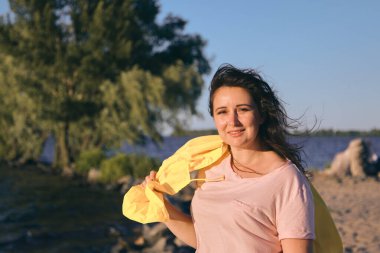 Geniş bir nehrin kumlu sahilinde uzun kızıl saçlı ve parlak sarı ceketli güzel bir kızın portresi.