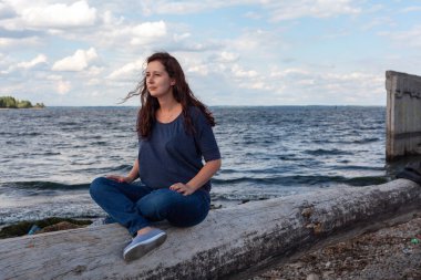 Genç tombul kadın nehrin kıyısındaki kütüğün üzerinde Lotus pozisyonunda oturuyor. Meditasyon ve doğayla bütünlük