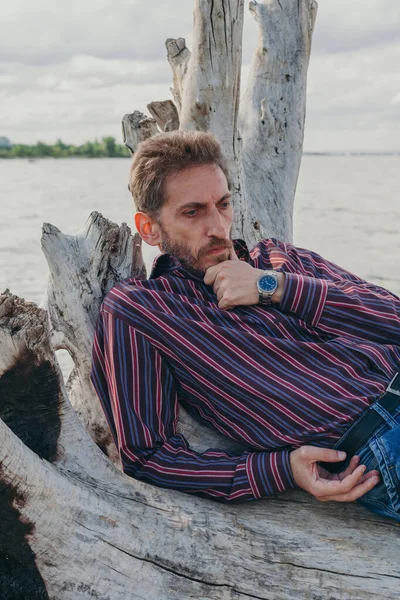 bearded man with a pensive look lies on an old driftwood log by the rive