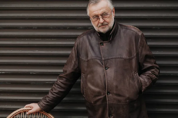 Portrait of a man with a gray beard. An elderly man in glasses and a leather jacket leans on a wicker chair. Rear Rolling Gate