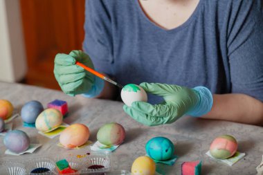 Kauçuk mavi eldivenli eller bir yumurta ve boya fırçası tutuyor. Ev yapımı Paskalya yumurtası ve renkli likörlü bardaklar.