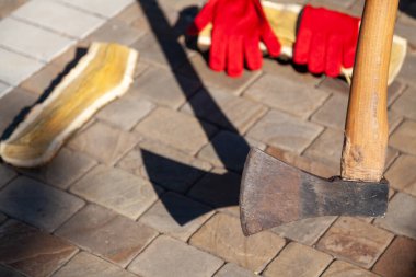 Balta, avludaki asfalt levhalara gölge düşürüyor. Yakınlarda doğranmış bir kütük ve kırmızı iş eldiveni var.