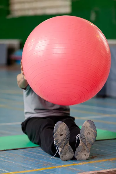 instructor sits on the floor of the gym and holds in front of him a large pink fitball for aerobic