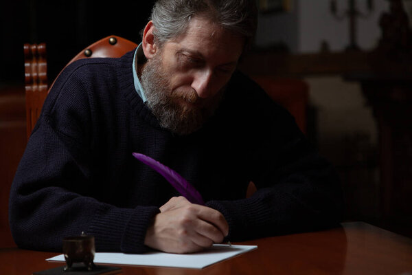 Человек с бородой сидит за большим полированным столом и пишет на белом листе гусиным пером и чернилами