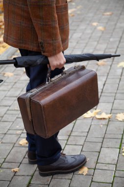 Şık bir ceketi olan beyefendi sonbahar öğleden sonra meydanın ortasında duruyor. Han 'ında eski bir çanta ve şemsiye kamışı tutuyor.