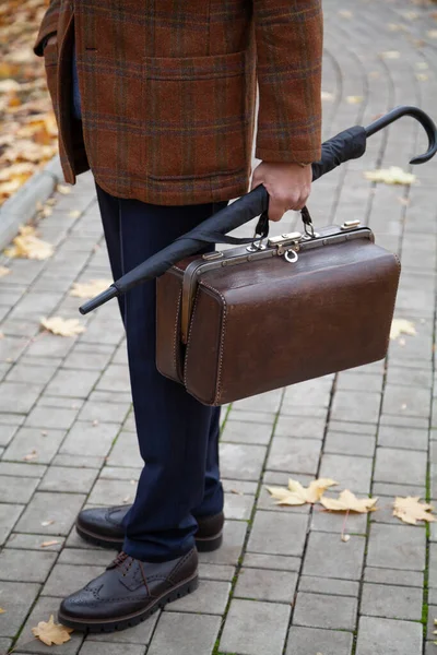 gentleman in a stylish checkered jacket stands in the alley of the square in the fall afternoon. Holding a vintage bag and umbrella cane in his han