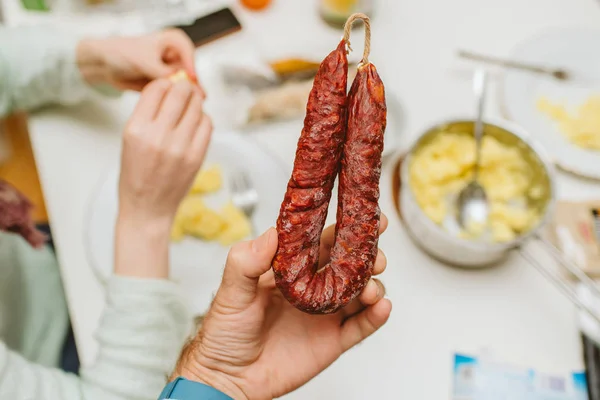 Man holding with dining table background a portion of Chorizo sausage with silhouette of woman eating in the background