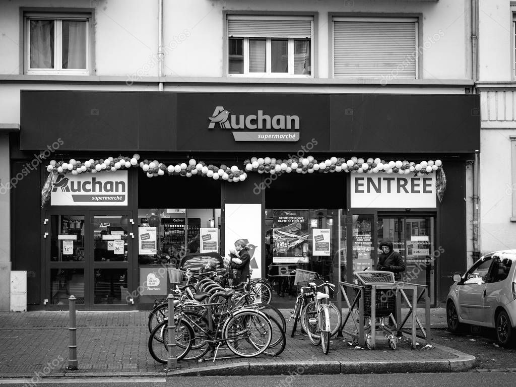 STRASBOURG, FRANCE - DEC 4, 2017: Black and white Auchan Supermarket entrance in French neighborhood on a winter snow day with customers exiting the entrance of the store