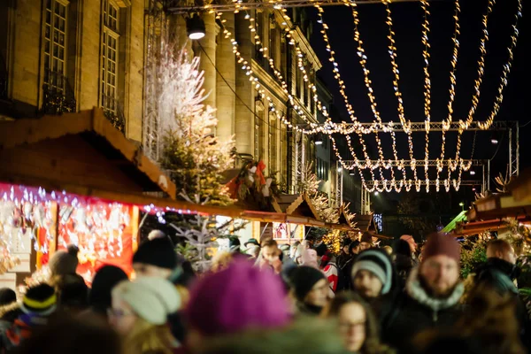 Velký dav lidí procházky pod vánoční světlo dekorace přes rušné ulici s tržními stánky a chaty prodávající dárky — Stock fotografie