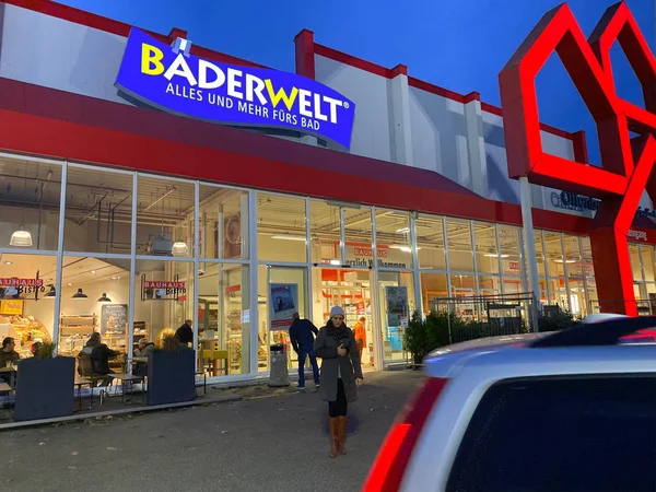 Широкий обзор немецкого строительного магазина Bauhaus с клиенткой, выходящей из магазина в сумерках и логотипом Badewelt — стоковое фото