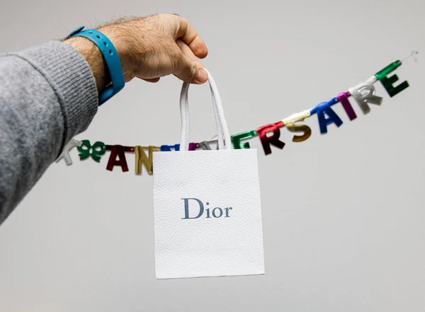 Man hand memegang hadiah baru dari Christian Dior kantong kertas putih melawan stiker Joyeux anniversaire di dinding — Stok Foto