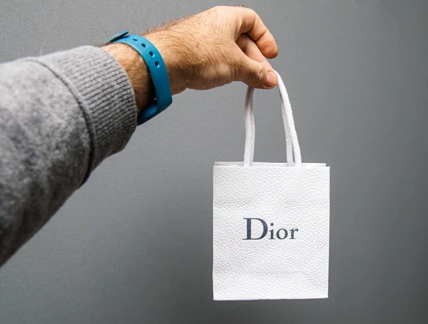 Mano del hombre sosteniendo el nuevo regalo de Christian Dior bolsa de papel blanco contra fondo gris — Foto de Stock