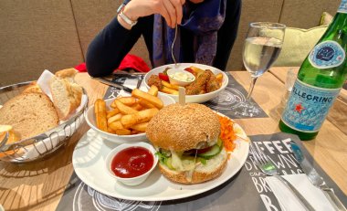 Lezzetli balık ve patates kızartması yiyen bir kadın. Yanında hamburger ve patates kızartması var. San Pellegrino maden suyu içiyor.
