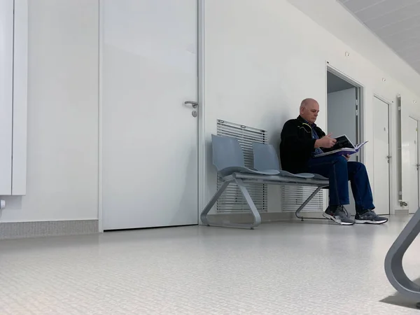 Curioso hombre mayor lectura resultados en el mapa dentro de la clínica moderna sala de espera hospital sentado en el banco gran corredor de perspectiva larga —  Fotos de Stock