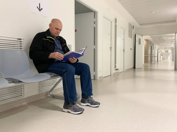 Senior man läser resultat i kartan inne i moderna kliniken sjukhus sitter på bänken stora lång perspektiv korridor — Stockfoto