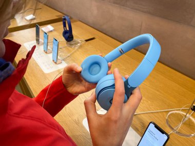 Apple Bilgisayar Mağazası 'ndaki kırmızı ceketli kadın, Dr. Dre Solo Pro Aktif Gürültüyü İptal Eden Kulaklıkları hayranlıkla dinliyor.