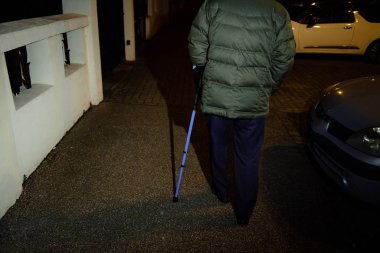 Gece Fransız caddesinde yürüyen yaşlı bir adamın silueti mavi teleskopik alüminyum kamış ile dengeyi koruyan baston kullanarak tıbbi yardım ve rehabilitasyon
