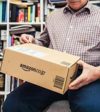 Paris, France - Apr 2, 2019: Senior holding new parcel arrived from Amazon Japan with large logotype on side and bibliotehque furniture in background