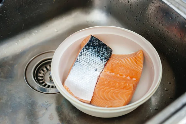 Kitchen stainless steel sink with plastic bowl containing two pieces of salmon — Stock Photo, Image
