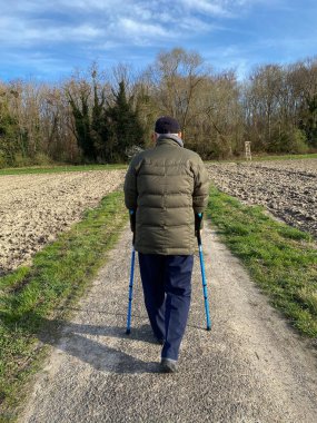 Yaşlı, kendine güvenen bir erkeğin kırsal yolda yürüyen tıbbi bastonlarla ormanda temiz havaya doğru yürüdüğünü görüyor.
