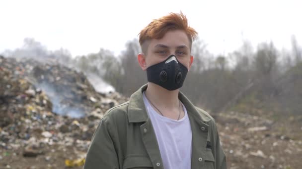 의료 마스크를 쓴 젊은 남성의 초상화가 거대 한 플라스틱 매립지 배경에서 카메라를 바라봅니다. 생태계 의대 재난 — 비디오