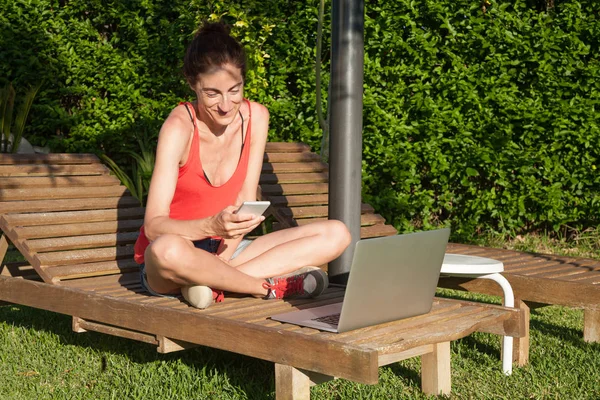 Mujer con teléfono inteligente y portátil en la silla de la cubierta del salón — Foto de Stock