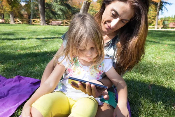 Маленькая девочка и мама смотрят мобильный телефон, сидя на траве — стоковое фото