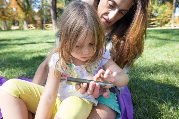 Little girl touching mobile screen of woman sitting in park — Stock Photo, Image