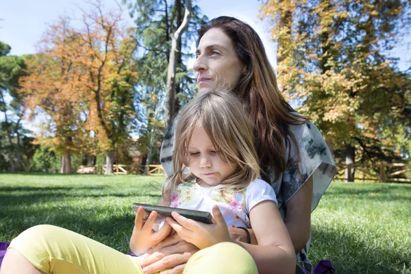 Маленькая девочка смотрит мобильный телефон сидя на ногах женщины в парке — стоковое фото