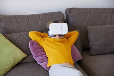 6 yaşındaki bir çocuk kahverengi bir koltukta rahatça oturuyor ve sanal gerçeklik kulaklığı takıyor.