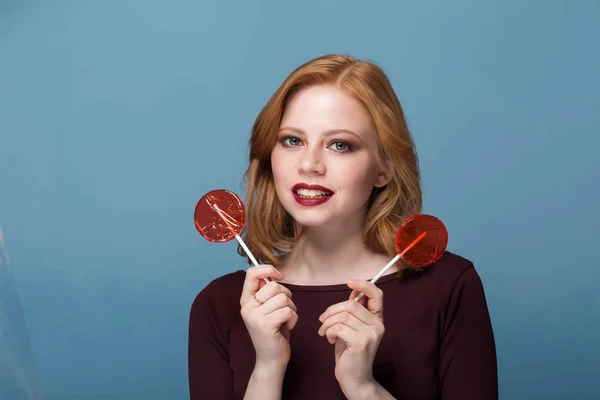 Beautiful redhead woman with two red lollipops on a blue background looks with a smile — Stock Photo, Image