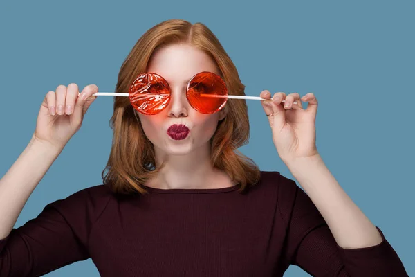 Menina bonita com dois doces vermelhos redondos fecha os olhos, parafusando seus lábios contornos rosto olhando para cima no fundo azul — Fotografia de Stock