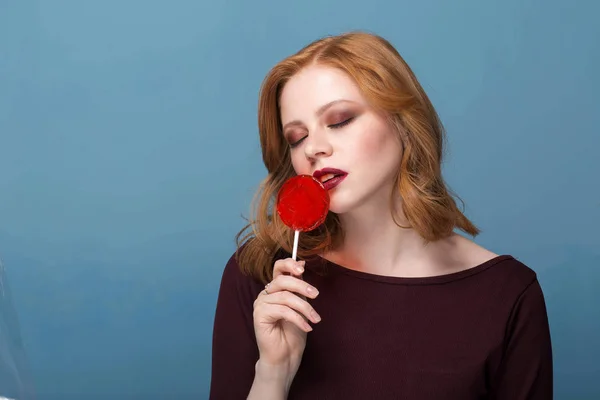 Modelo feminino elegante posando com pirulito e com olhos fechados no fundo azul . — Fotografia de Stock