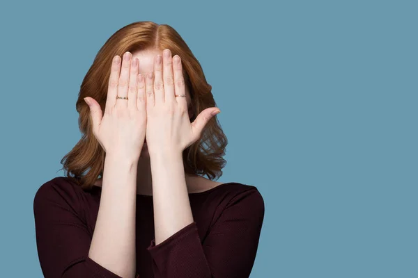 Kızıl saçlı kız bordo ceketli bir mavi arka plan ayrı tut moduyla boşaltmak duyguları üzerinde iki elleriyle yüzünü kapsar