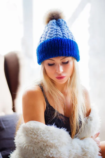 Sensitive blonde woman looking down. Girl wearing a blue knitted hat and a white coat.