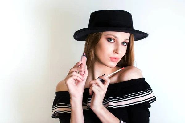 Hermosa mujer rubia, blogger de belleza con sombrero negro tiene lápiz labial en la mano. Cosméticos, brillo de labios, cuidado de la cara, maquillaje . — Foto de Stock
