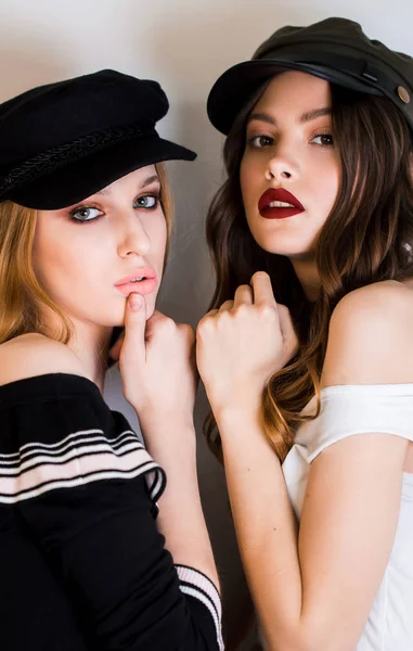 Deux belles femmes, filles blondes et brunes en casquettes noires et maquillage lumineux regardent la caméra — Photo