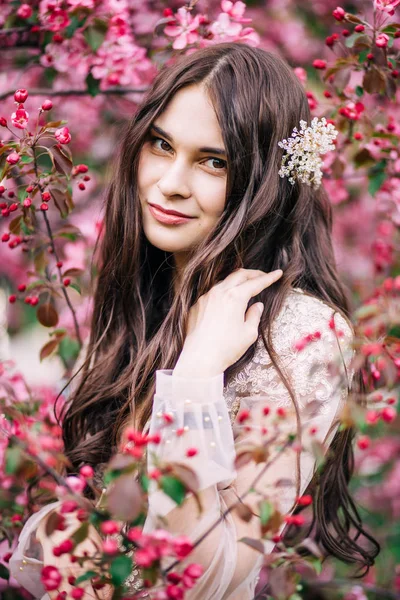 Belle jeune mariée mignonne en dentelle de boudoir robe transparente, avec décoration sur les cheveux, souriant, regardant autour de l'arbre fleurit avec des fleurs roses — Photo