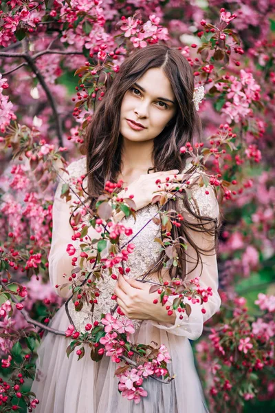 Belle fille dans une robe se tient près de l'arbre fleurit avec des fleurs roses, tenant par la main un brin, lève les yeux avec un sourire — Photo