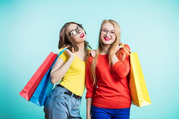 Twee gelukkige jonge vrouwen met boodschappentassen op blauwe achtergrond kijk met een glimlach. Verkoop, shopping, toerisme en gelukkige mensen. — Stockfoto