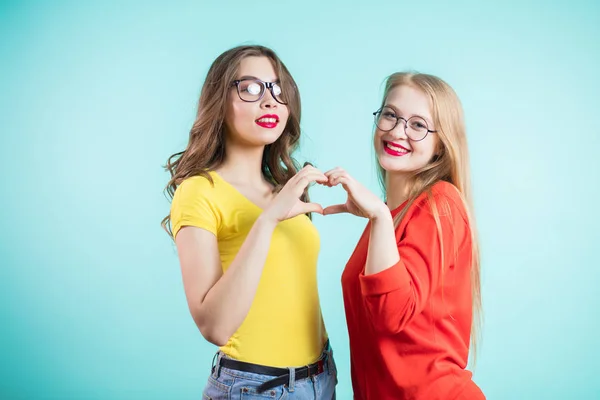 Twee mooie jonge vrouwen tonen duimen hart en met een glimlach kijken naar de camera op een blauwe achtergrond — Stockfoto