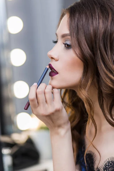 Linda chica linda en un vestido azul pintado en el espejo, poniendo en lápiz labial maquillaje profesional, maquillaje, perfil, primer plano — Foto de Stock