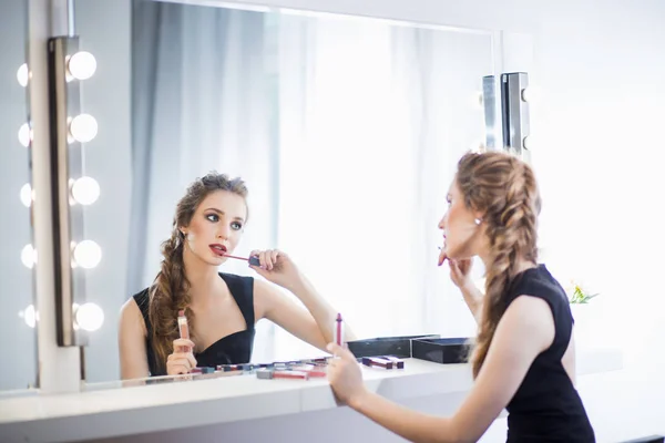 Piękna dziewczyna siedzi w tabeli make-up artist w pobliżu lustra, stosowane szminki, lip gloss, Studio, salon, kosmetyki — Zdjęcie stockowe