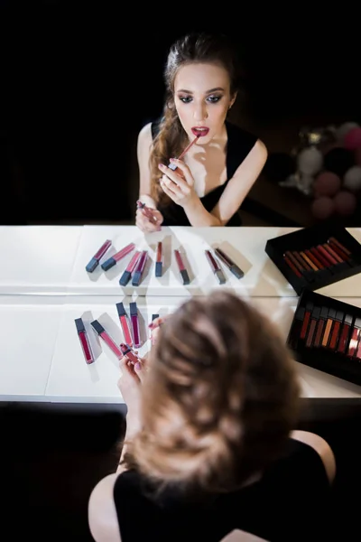 Gadis cantik berambut pirang seksi duduk di meja make-up artis di dekat cermin, menerapkan lipstik, lip gloss, Studio, salon, makeup, latar belakang hitam — Stok Foto