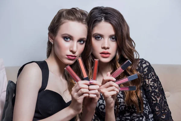 Dos hermosas chicas en vestidos negros con maquillaje profesional, peinado, sentado en el sofá, anunciar cosméticos, están sosteniendo una gran cantidad de lápices labiales, brillo de labios — Foto de Stock